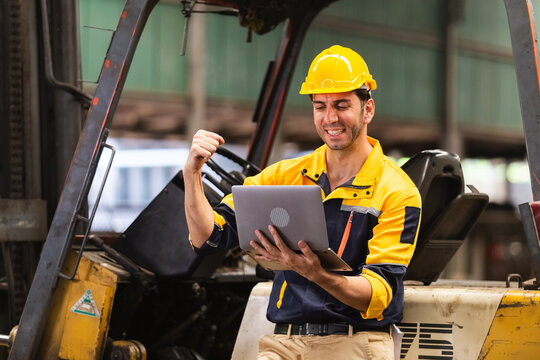 Happy industrial worker in safety uniform celebrates success while using laptop near forklift in factory. Concept of digital transformation and efficiency improvement in manufacturing.