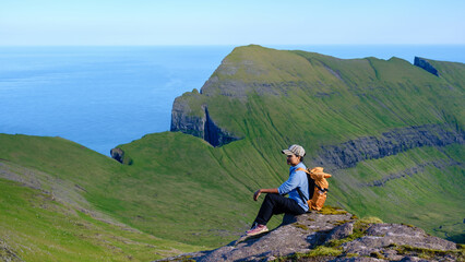 A solitary hiker takes in the expansive view from a rocky ledge in the Faroe Islands, surrounded by...