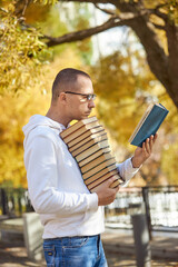 Fototapeta premium A young man stands in a park, enjoying a sunny afternoon. He is holding a book in one hand while balancing a tall stack of books in the other. Gorgeous autumn leaves surround him