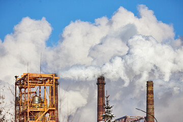 Factory structures release visible smoke plumes while standing against a backdrop of blue sky and...
