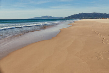 Quiet sandy beach stretches along the coastline, with gentle waves lapping at the shore and distant mountains creating a serene backdrop under clear blue skies