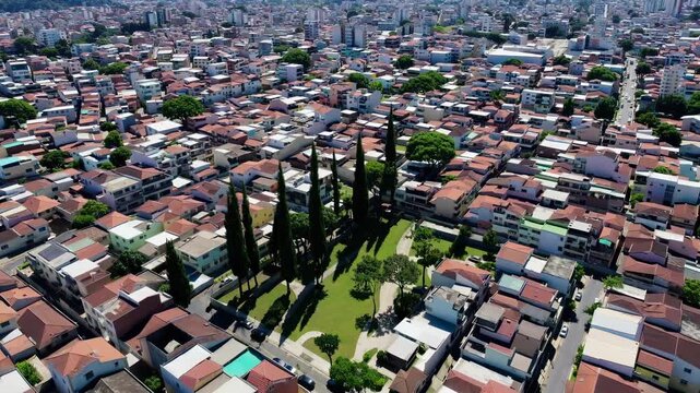 Aerial view of a densely populated urban area with numerous red-roofed houses and scattered tall trees. A small park area is visible in the midst of the residential structures.