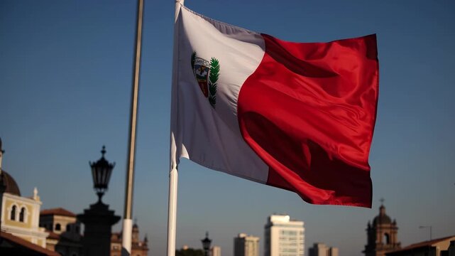 The national flag of Peru with its coat of arms is blowing in the wind against a clear blue sky. The background includes a few distant structures and a street lamp silhouette.