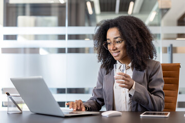 Fototapeta premium An African American businesswoman works in the office at a laptop and holds a glass of clean water in her hand