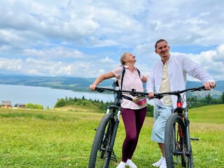 Fototapeta premium Happy smiling couple with bicycles on a cycling path near Czorsztyn and Czorsztynskie Lake with Tatra Mountains in the background, Poland. Concept of active lifestyle, love, adventure, and eco tourism