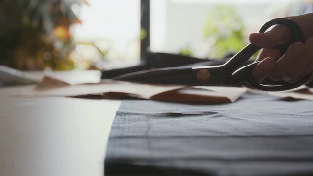 Close-up of hand of anonymous seamstress carefully cutting dress or trousers pattern from black cloth with sharp scissors, while creating designs in artisan workshop
