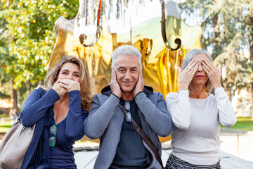 Three male and female friends are sitting in front of a large golden abstract sculpture replicating the gesture of the three wise monkeys