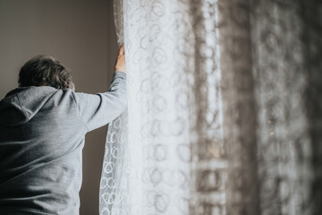 Elderly individual looks outward through sheer white curtains, evoking feelings of contemplation...