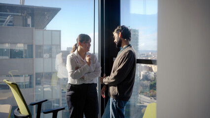 Two colleagues standing by a large office window having a casual conversation in natural light, representing workplace communication, connection and collaboration.