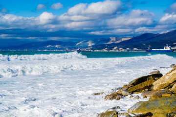 Obraz premium Kabardinka resort. Large green storm waves in white foam roll over rocky beach of Kabardinka. In background is port of Novorossiysk and Caucasus Mountains. Large stones on shore. 