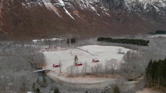 An aerial view captures the serene beauty of a snow dusted valley near Trollstigen, Norway, featuring traditional red cabins nestled amongst frosted trees and a winding river against a backdrop of maj