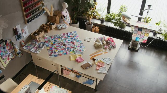 Full no people top view of cutting table at craft workshop or manufactory, with patchwork garment, scraps of fabric, rail with handmade clothes and merchandise, steam ironing station by large window