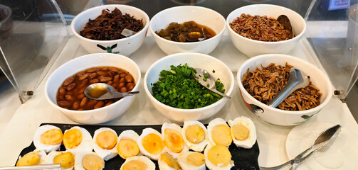 A selection of savory toppings for porridge, including braised beans, chopped scallions, fried shallots, and salted egg halves.