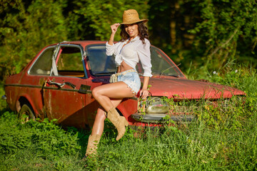 Beautiful woman with a straw hat poses next to an old abandoned car surrounded by vegetation.