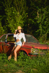 Beautiful woman with a straw hat poses next to an old abandoned car surrounded by vegetation.