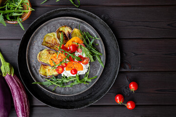 Salad with fried eggplant, cream cheese, tomatoes and arugula on black wooden background top view. Vegetarian food