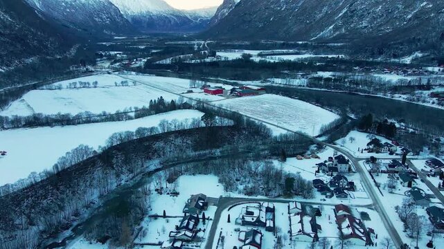 An aerial drone shot captures the serene winter landscape of Oppdal, Norway, showcasing snow covered fields, a winding river, and charming houses nestled within the valley, surrounded by majestic moun