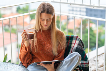 Relaxed young woman sitting on balcony wrapped in blanket, holding smartphone with cup of coffee nearby. Concept of communication, social media and mindful morning