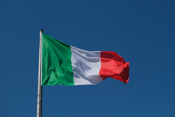 The Italian tricolor flag, with its green, white, and red stripes, waves gracefully in the wind against a clear and bright blue sky.