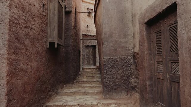 Narrow alley in a Moroccan village with stone steps and wooden doors and window shutters. The walls are made of mud bricks. The scene evokes a sense of history and tradition.