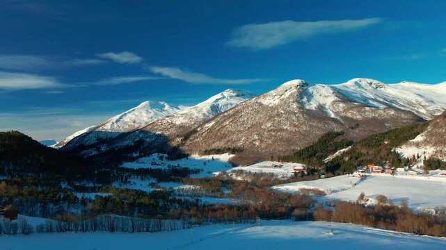 aerial view captures the serene winter landscape of Oppdal, Norway. Snow capped mountains frame a picturesque valley dotted with charming traditional houses, surrounded by pristine forests and fields 