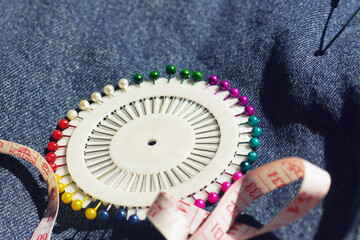 Rainbow pin wheel with colorful ball head pins and measuring tape on denim background. Educational craft supplies for learning basic tailoring skills, sewing classes, textile arts curriculum programs.