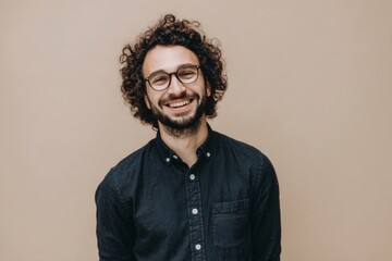 Obraz premium Friendly smiling man with curly hair and glasses wearing a dark shirt against a beige background.