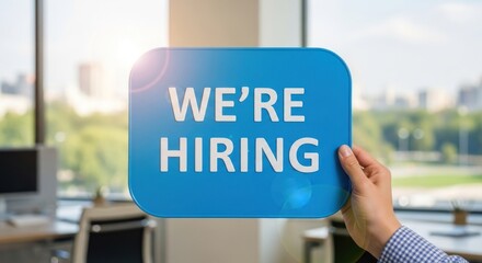 Hand holds "We're Hiring" sign, city view background, office setting