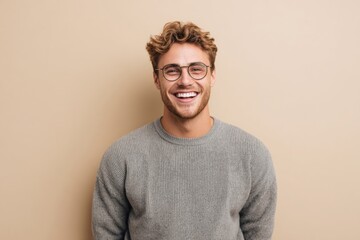 Smiling young man with glasses and curly hair wearing casual sweater standing against beige background.