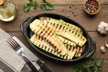 Delicious grilled courgette slices in baking dish, oil and spices on wooden table, flat lay