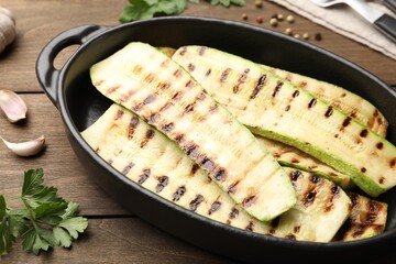 Delicious grilled courgette slices in baking dish and parsley on wooden table, closeup