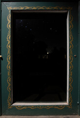 A mysterious night-time reflection in a dark window, framed by an elegant dark green and ornate gold border that adds a touch of classic, moody glamour.
