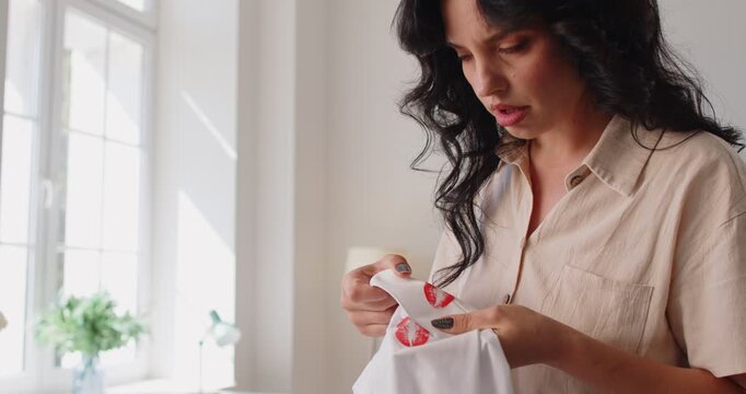 Lipstick stains on shirt alarm woman. She checks the collar, spotting kiss marks and infidelity. Daylight fills a tidy home while suspicion builds. Concept: trust, doubt, betrayal in love.