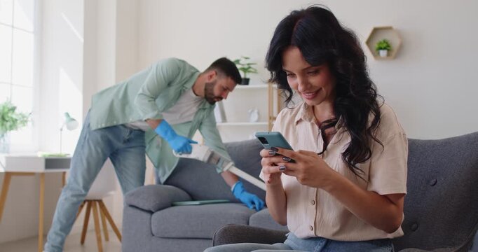 Woman secretly texts on her phone while her partner cleans, showing mistrust and hidden infidelity suspicion. Couple trust issues theme. A scene of emotional distance and betrayal.
