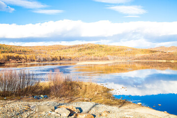 Fototapeta premium Calm lake reflecting bright sky and colorful autumn trees. Beautiful fall nature landscape scene for travel or seasonal design.
