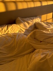 Messy bed with warm sunlight casting shadows across white pillows and sheets in the morning. Concept of rest, real life, slow living, comfort, and cozy domestic atmosphere.