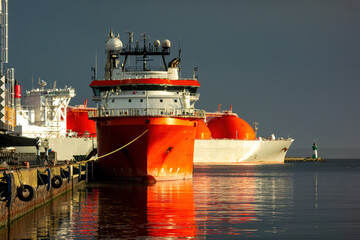 Industrial port scene with a red offshore supply vessel and an LNG tanker moored in the harbor. Symbolizing the global energy industry, logistics, and maritime transport.