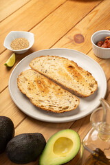 A plate with two slices of toasted bread surrounded by sesame seeds, dried tomatoes, avocados and olive oil.
