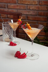 Two spicy dirty martinis served in a martini glass on a white table. It is garnished with olives and a red hot chilli pepper slice. A bricked wall is the background