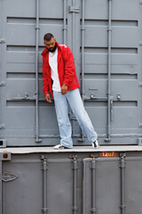 African american man in red windbreaker jacket posing on cargo containers in port  © Dmytro Hai