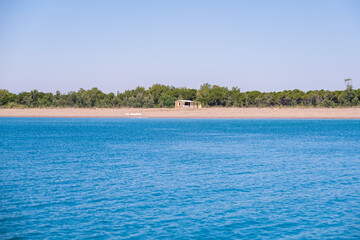 A blue sea with a sandy beach and a small shed in the distance, surrounded by green trees. Summer scene for travel.