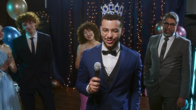 Happy prom king, wearing cown and matching navy bluesuit giving speech into mirophone at high school prom party