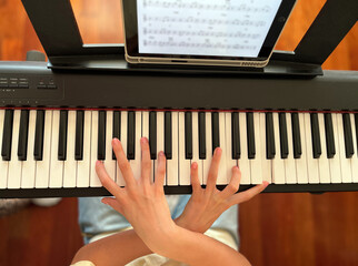  Teenager practicing piano at home with headphones, using a tablet app for online music lessons. Concept of digital education, e-learning. Close up view of hand and piano.