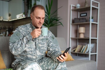 Soldier relaxing on sofa, drinking coffee and using smartphone