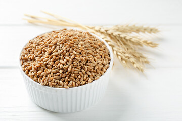Wheat grains in bowl and spikelets on white wooden table, closeup. Space for text