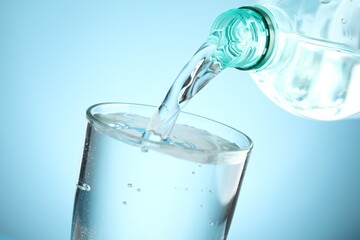 Pouring refreshing soda water from bottle into glass at table against light blue gradient background, closeup