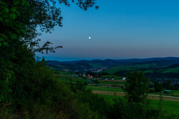 Obraz premium Picturesque rural valley with fields, houses, and winding roads under a rising full moon. The calm twilight atmosphere highlights harmony between village and nature.
