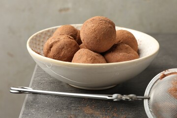 Tasty chocolate candies with cocoa powder in bowl on grey table, closeup