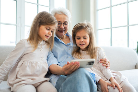 Happy family at home. Two little girls sisters twins grandmother enjoying time together watching video on phone. Good time at home. Grandma granddaughters child kids together. Family generations