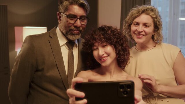 Young girl with curly hair, wearing pink fancy evening dress, taking selfies wih her parents in her bedrom, before going to high scool prom party